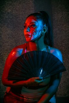 A striking portrait of a woman with artistic makeup, bathed in red and blue light, holding a fan.