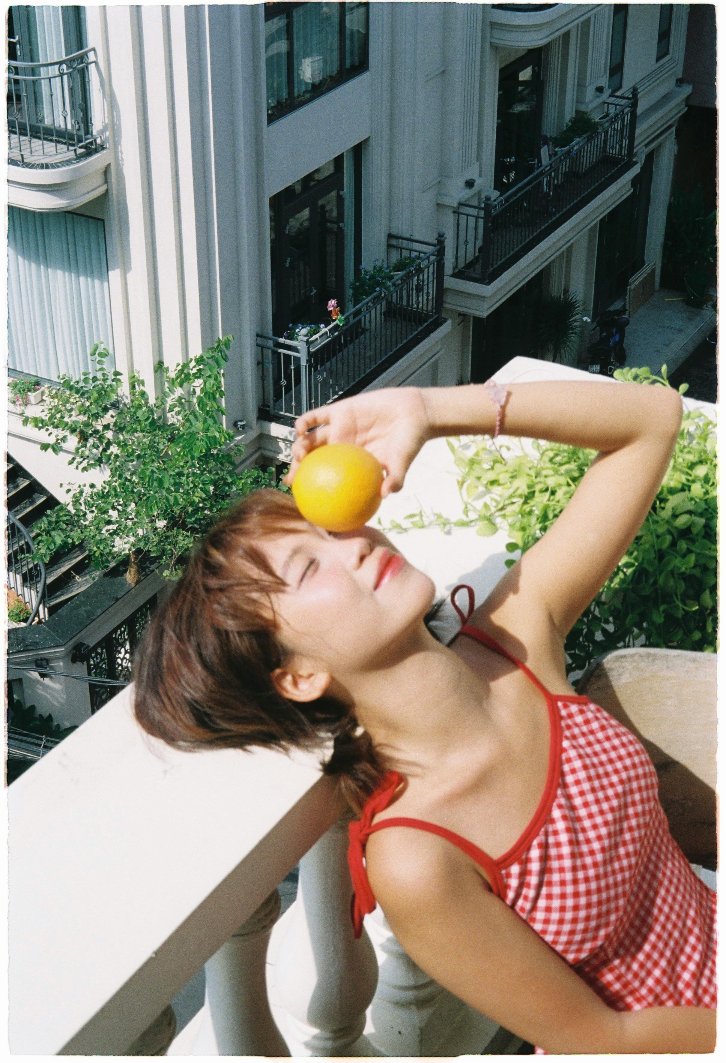 Relaxed woman enjoying sunlight with an orange on a balcony, embracing summer vibes.