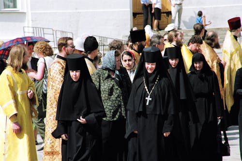 Nuns of convent wearing black clothes walking along street passing men of cloth in golden color robes and common people located near building celebrating holiday in sunny day
