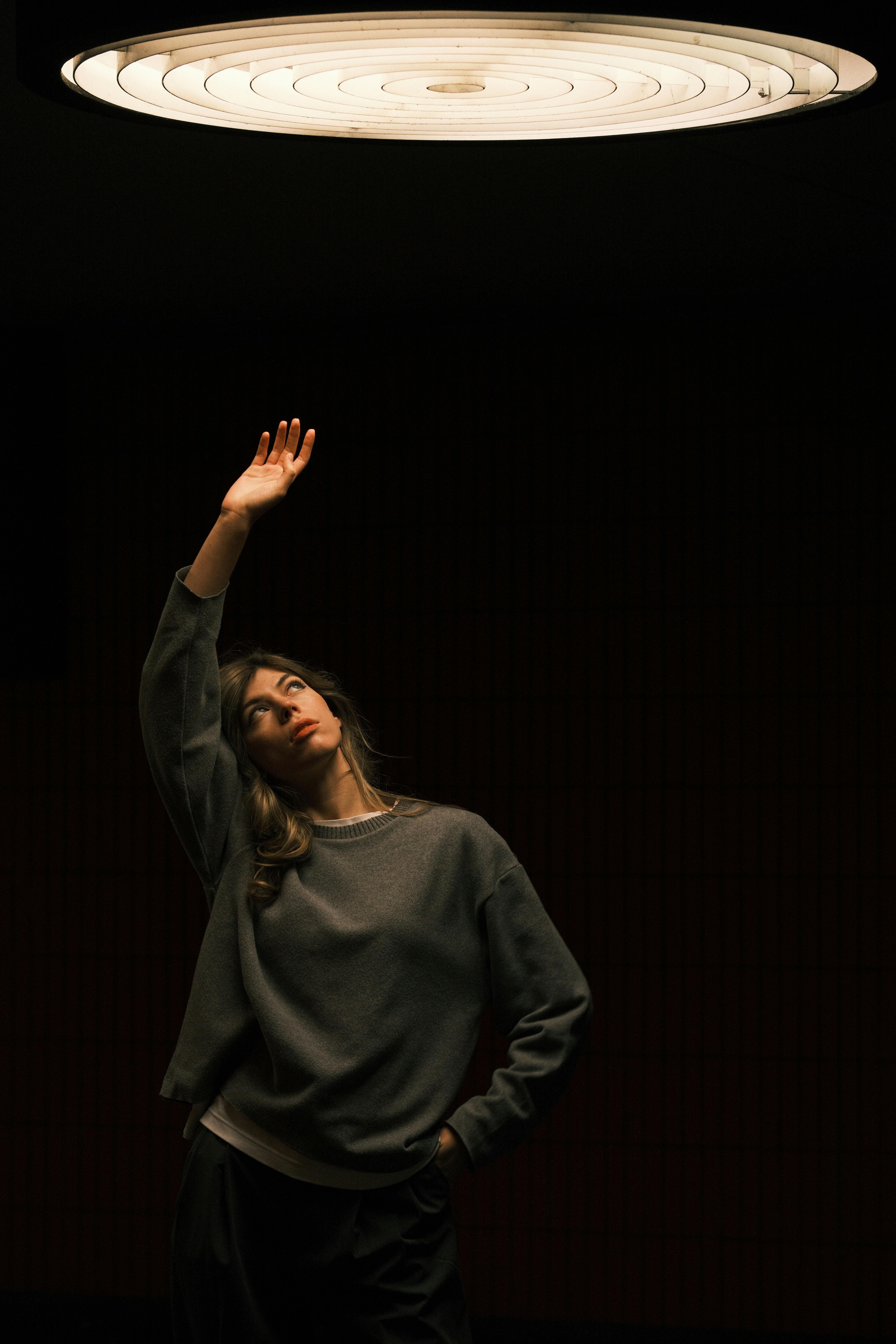 A woman stands under dim lighting, reaching towards a circular ceiling light.