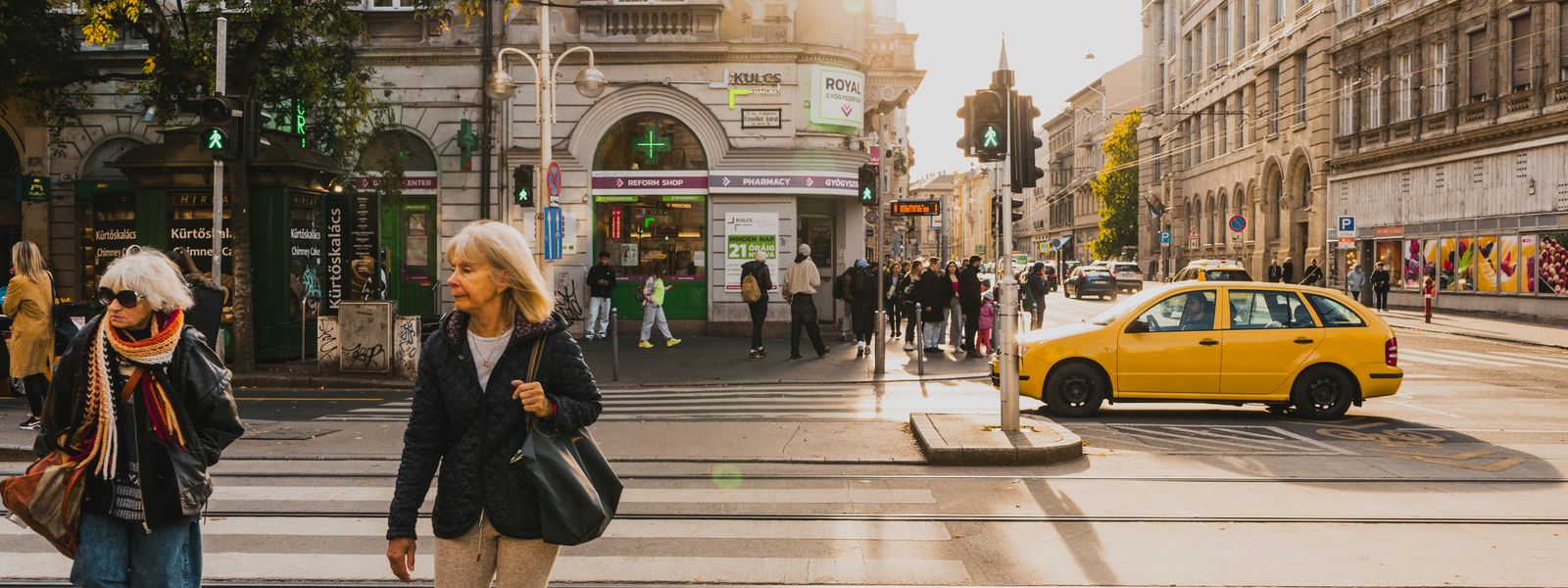 A bustling street intersection in Budapest with pedestrians and a yellow taxi under soft sunlight.