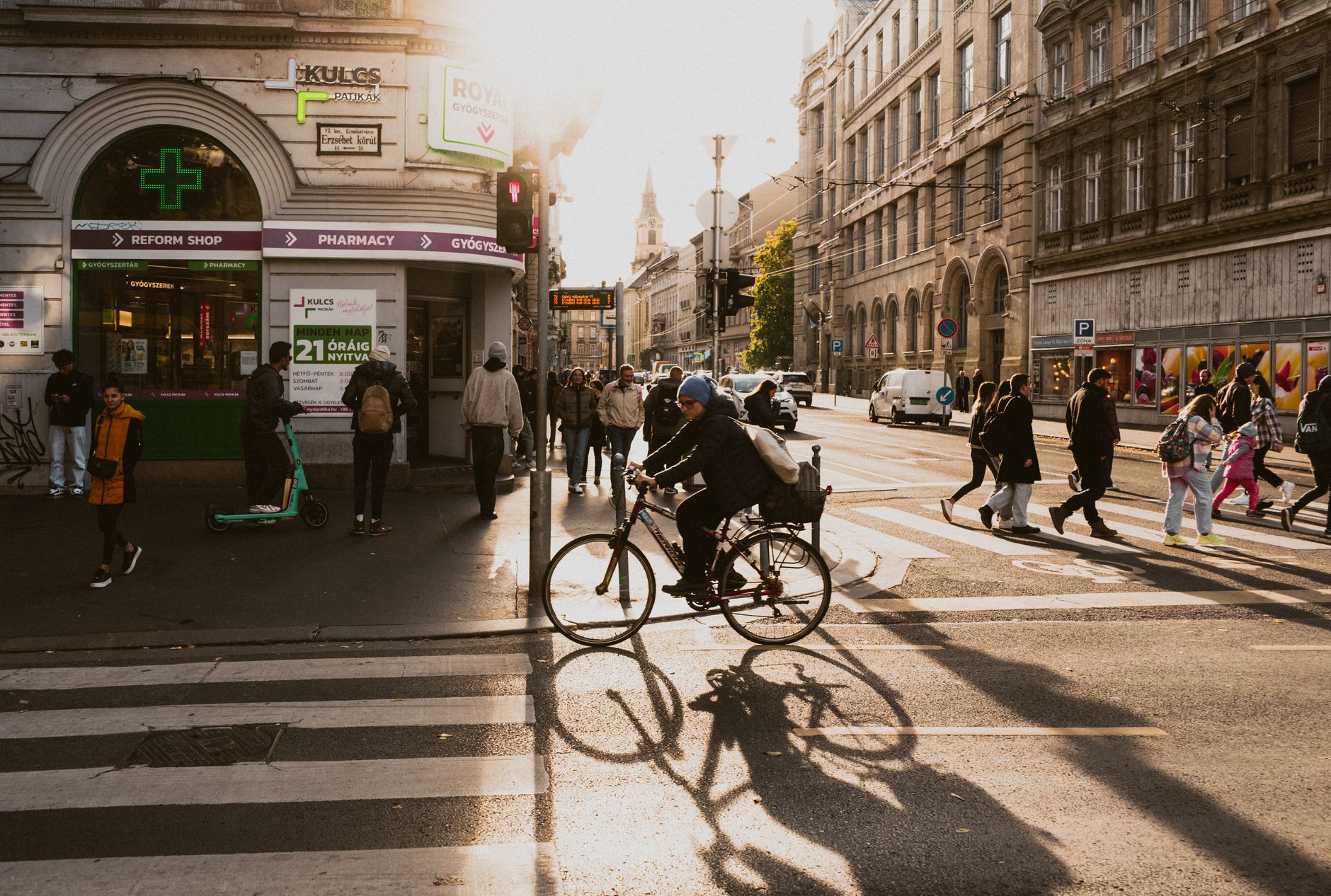 People crossing a sunlit street in Budapest with strong shadows.