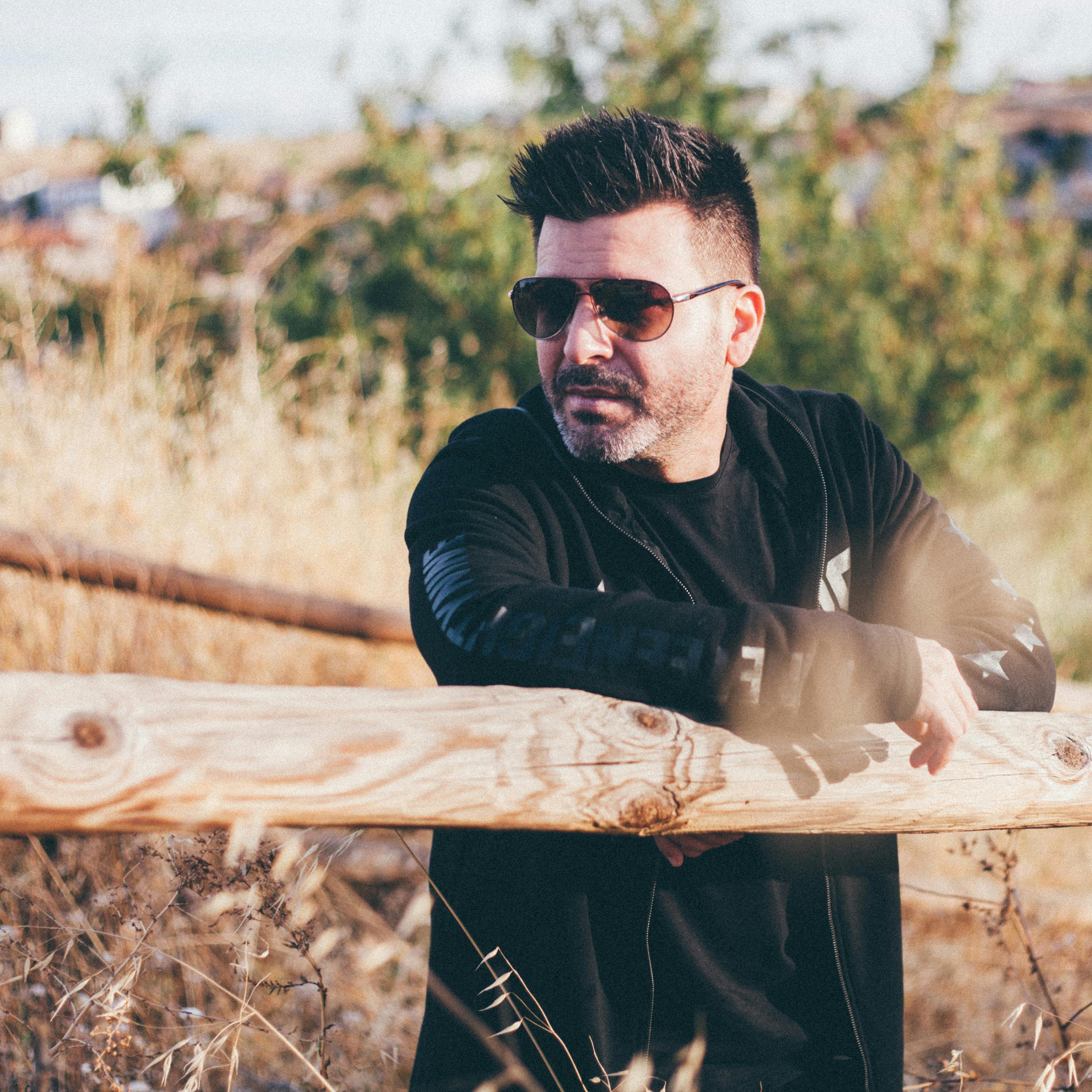 Adult male wearing sunglasses leans on a wooden fence in a sunny outdoor setting.