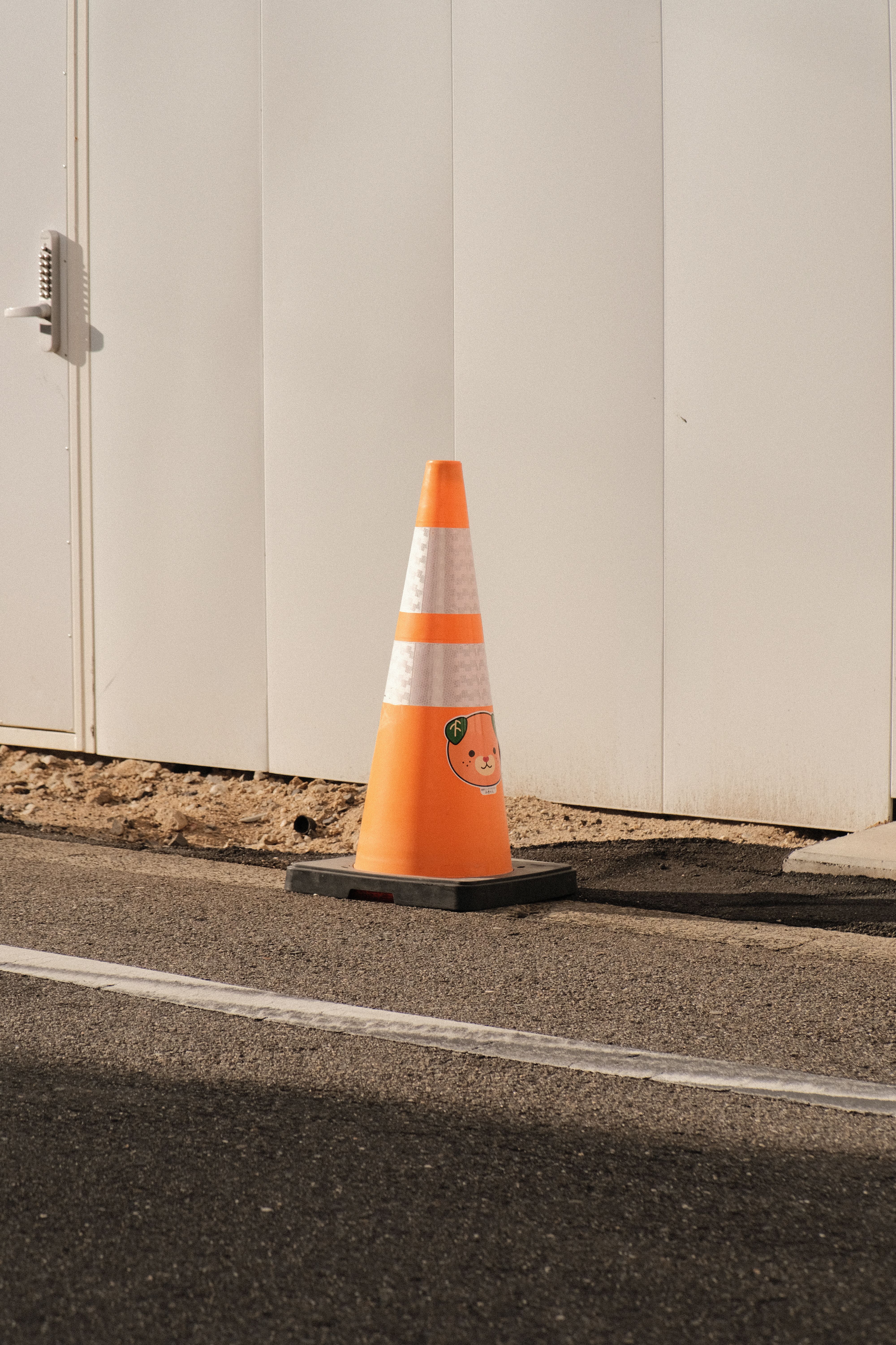A brightly colored traffic cone placed on a city street beside a white wall.