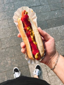 Hand holding a hot dog with ketchup and mustard on a city street.