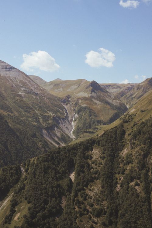 Free Stunning view of a mountain range with lush greenery and a waterfall under a clear blue sky. Stock Photo