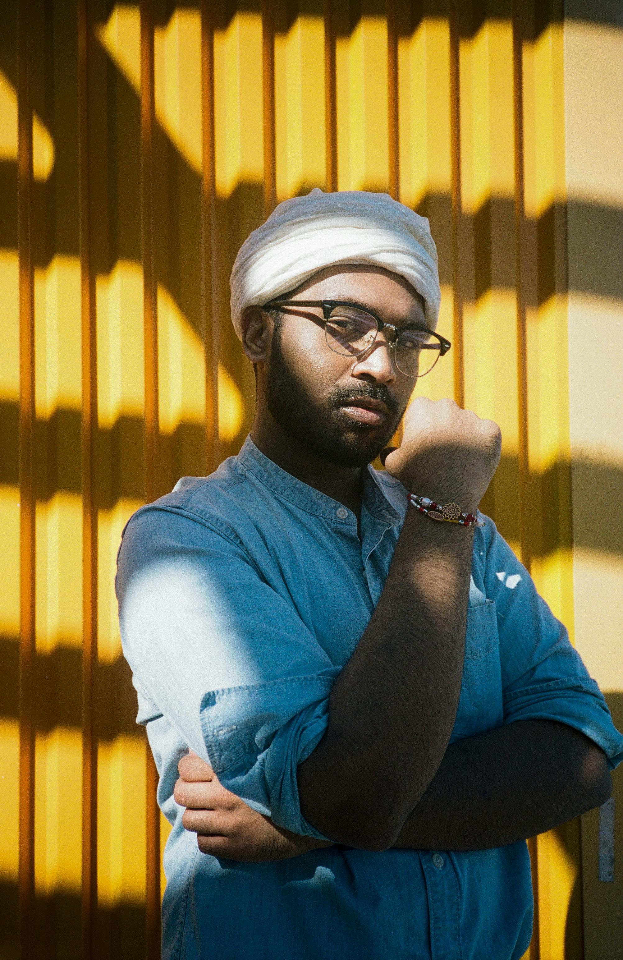 A stylish man in a turban poses confidently by a vibrant yellow wall under intense sunlight.