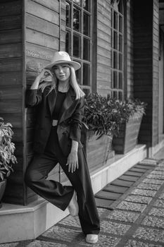 Elegant woman poses in black suit and hat, leaning on wooden wall.