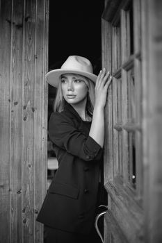 Black and white photo of a woman in a hat and suit standing at a rustic doorway, exuding sophistication.