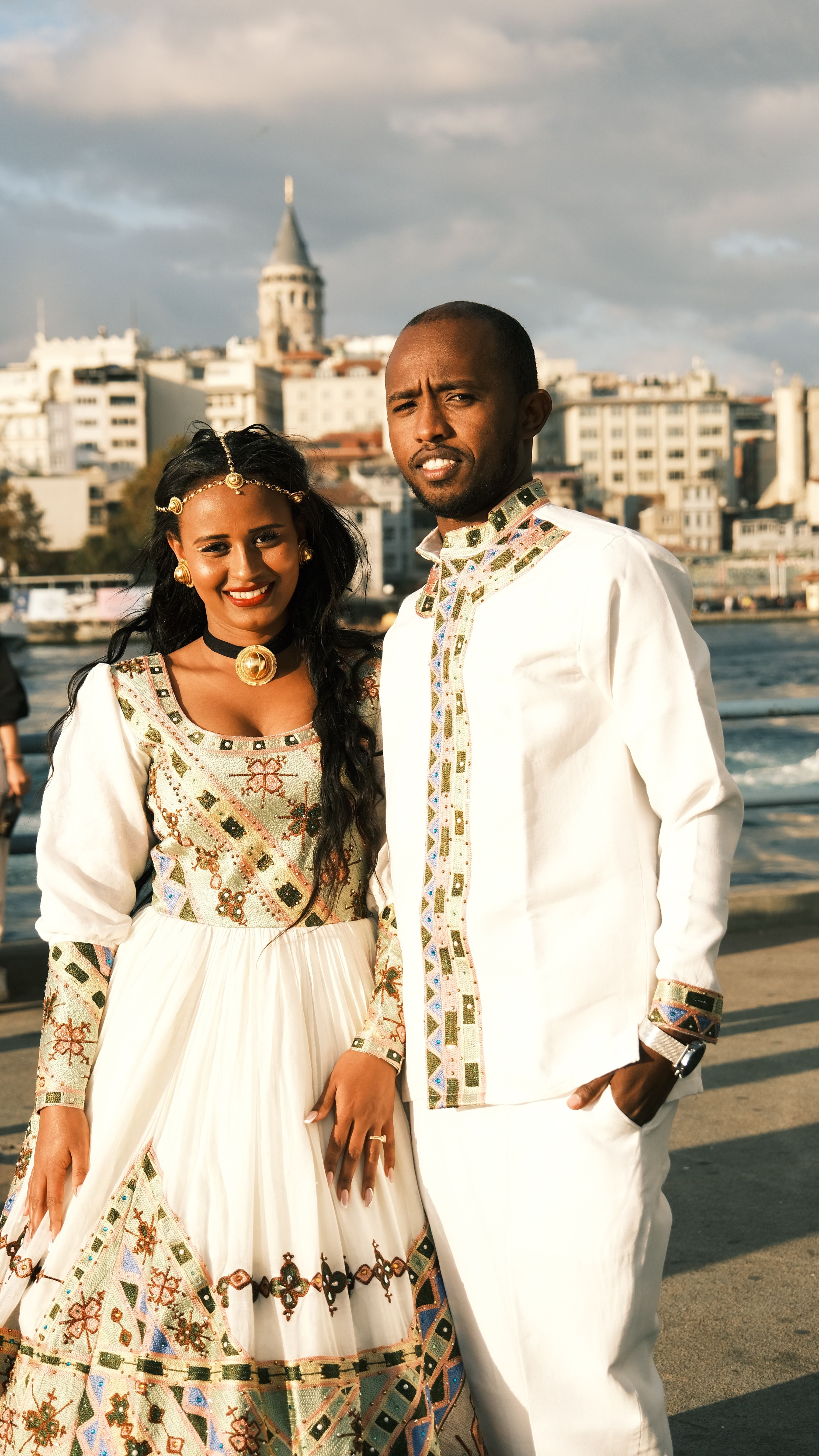 Couple in traditional attire posing by a waterfront with a city skyline.