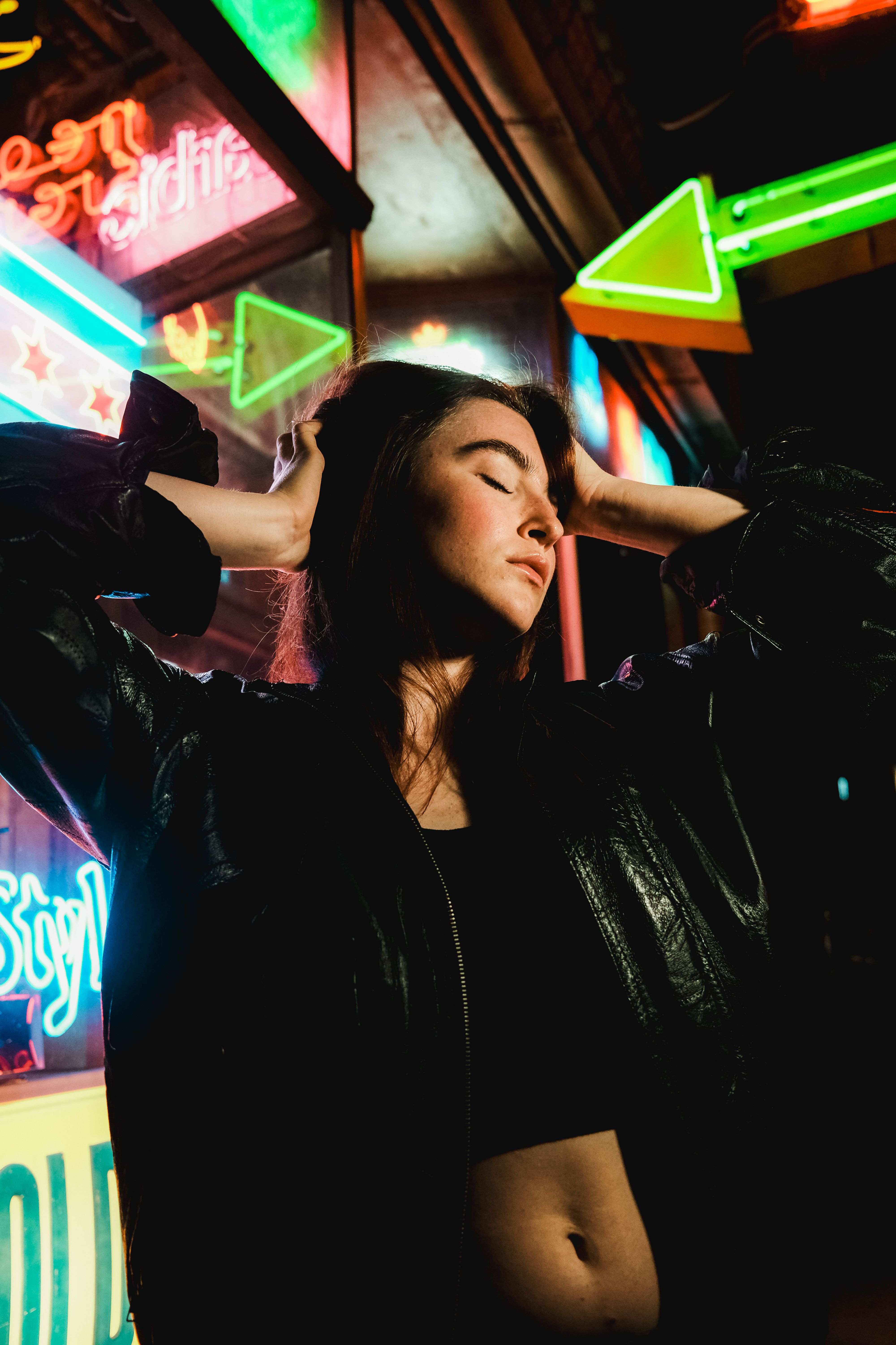 A young woman in a black jacket poses confidently in front of vibrant neon lights, capturing a modern city vibe.