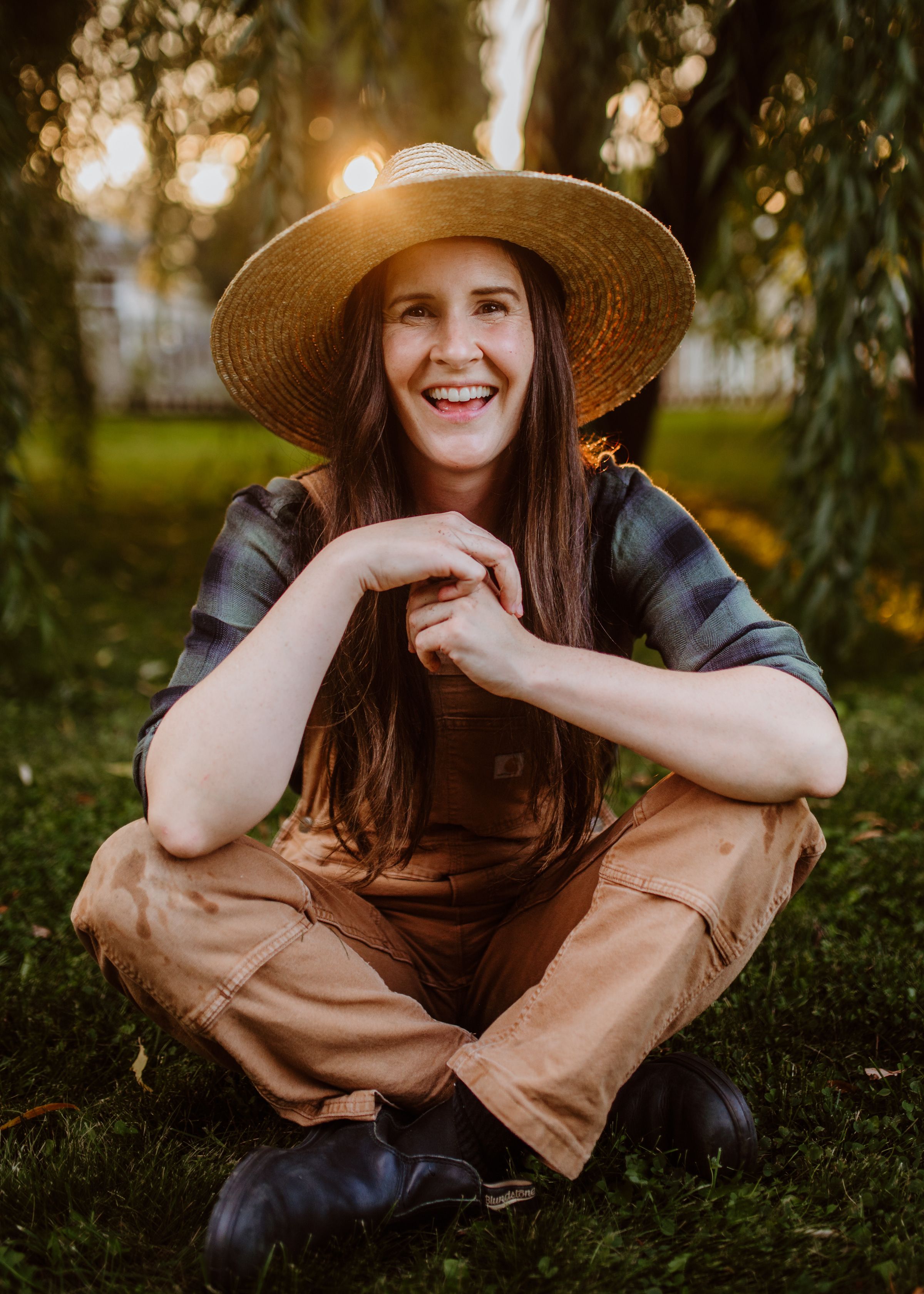 A woman in overalls and a sun hat smiles while sitting on grass outdoors, capturing a serene and joyful moment.
