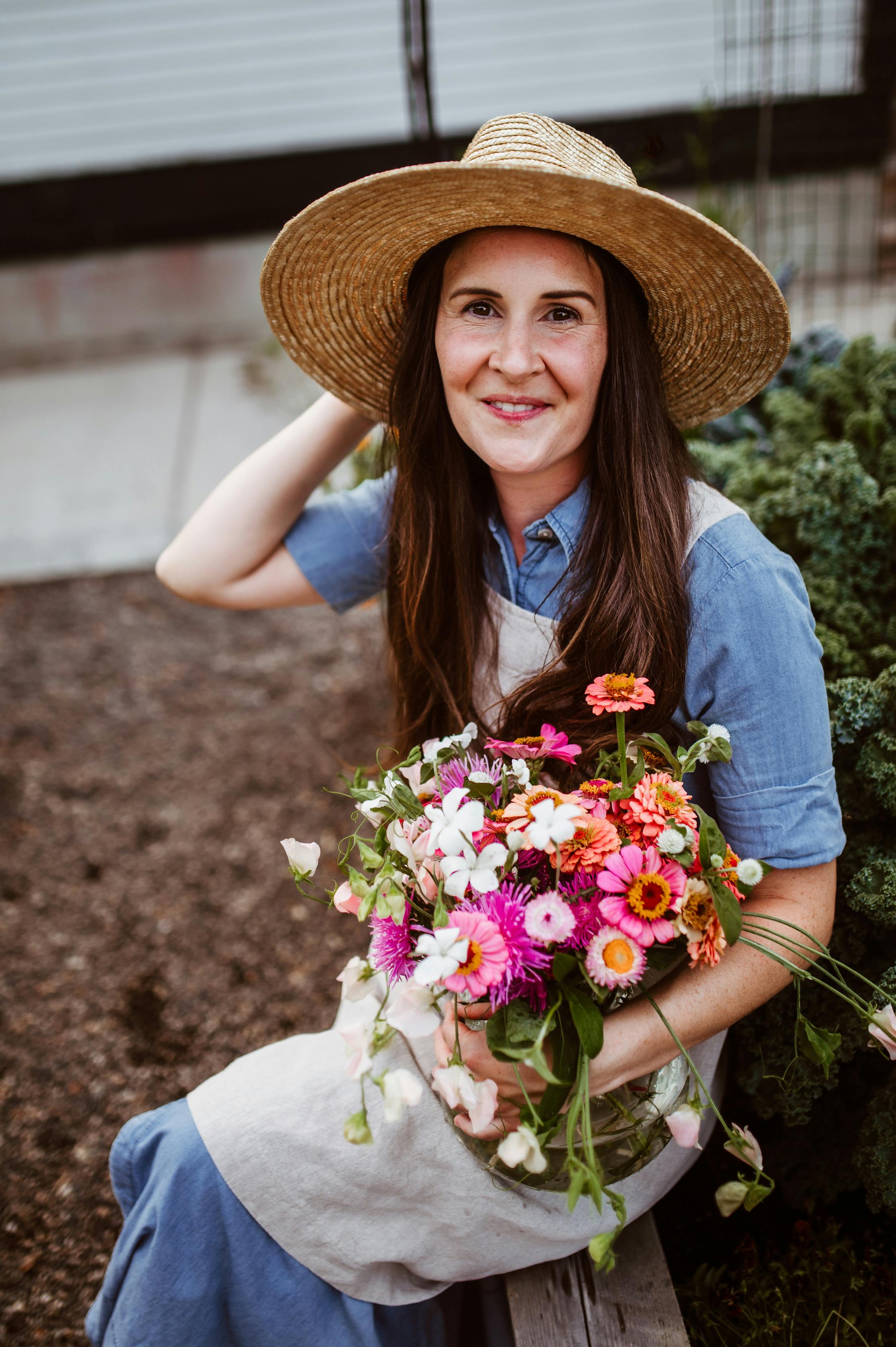 Woman in a straw hat sits happily in a garden holding colorful flowers.