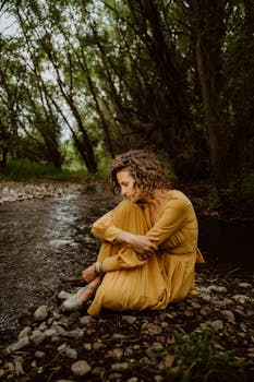 A woman in yellow sits pensively by a forest stream, reflecting amidst nature.