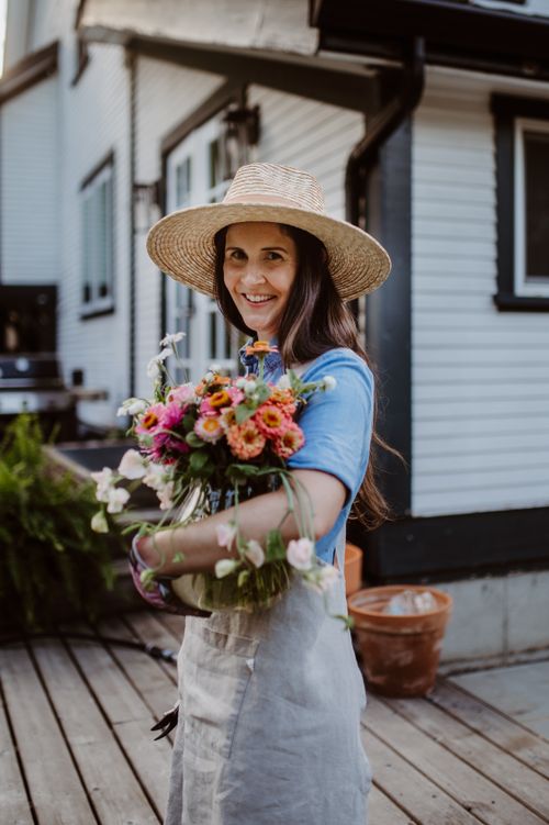 Free Woman in a straw hat holding vibrant flowers near a home garden, showcasing fresh blooms. Stock Photo