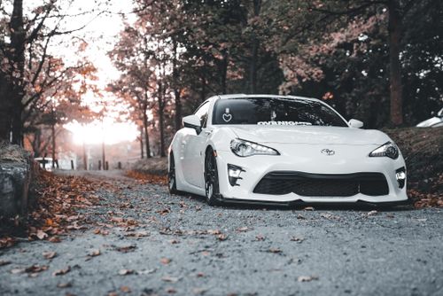 Free A sleek white sports car parked on a leaf-covered road in autumn scenery. Stock Photo