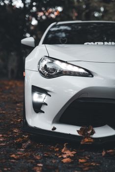 Close-up of a white sports car in an autumn forest setting, highlighting the headlight details.