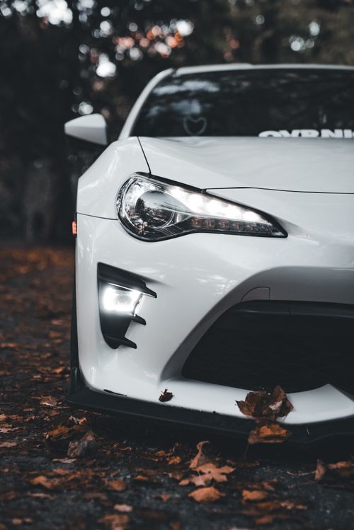 Free Close-up of a white sports car in an autumn forest setting, highlighting the headlight details. Stock Photo