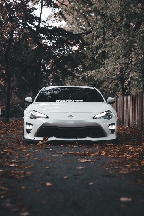 Free A sleek white sports car parked on a leaf-strewn road amidst autumn foliage. Stock Photo