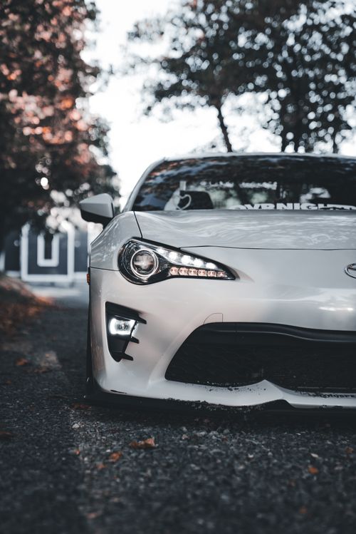 Free Dynamic shot of a white sports car parked on a rustic tree-lined street, highlighting sleek design. Stock Photo