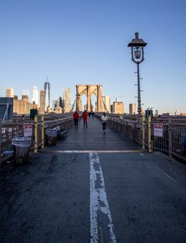 Pedestrians walking on the Brooklyn Bridge with New York City skyline in the background.