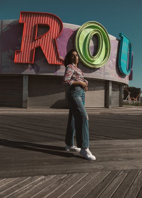 Stylish woman poses on Coney Island boardwalk with vibrant signage on a sunny day.