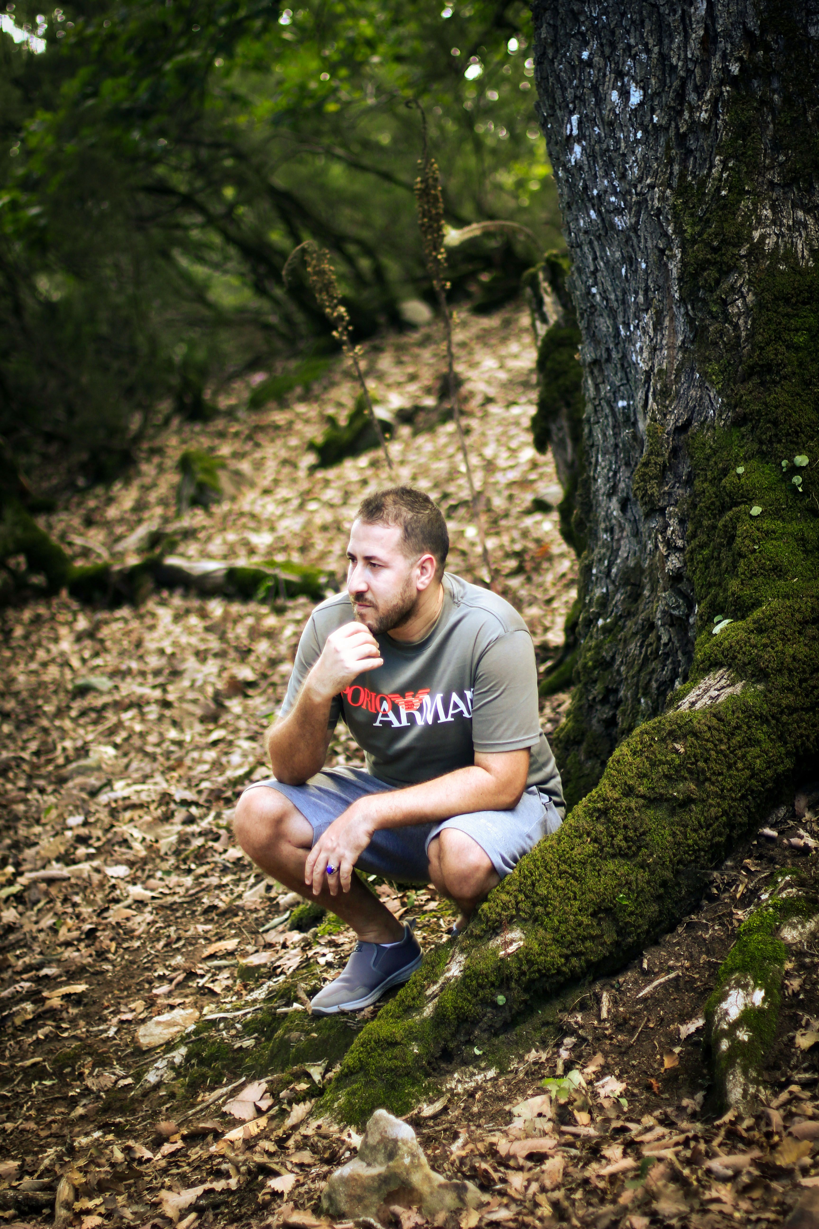 A thoughtful man squats beside a moss-covered tree in Skikda, Algeria forest.