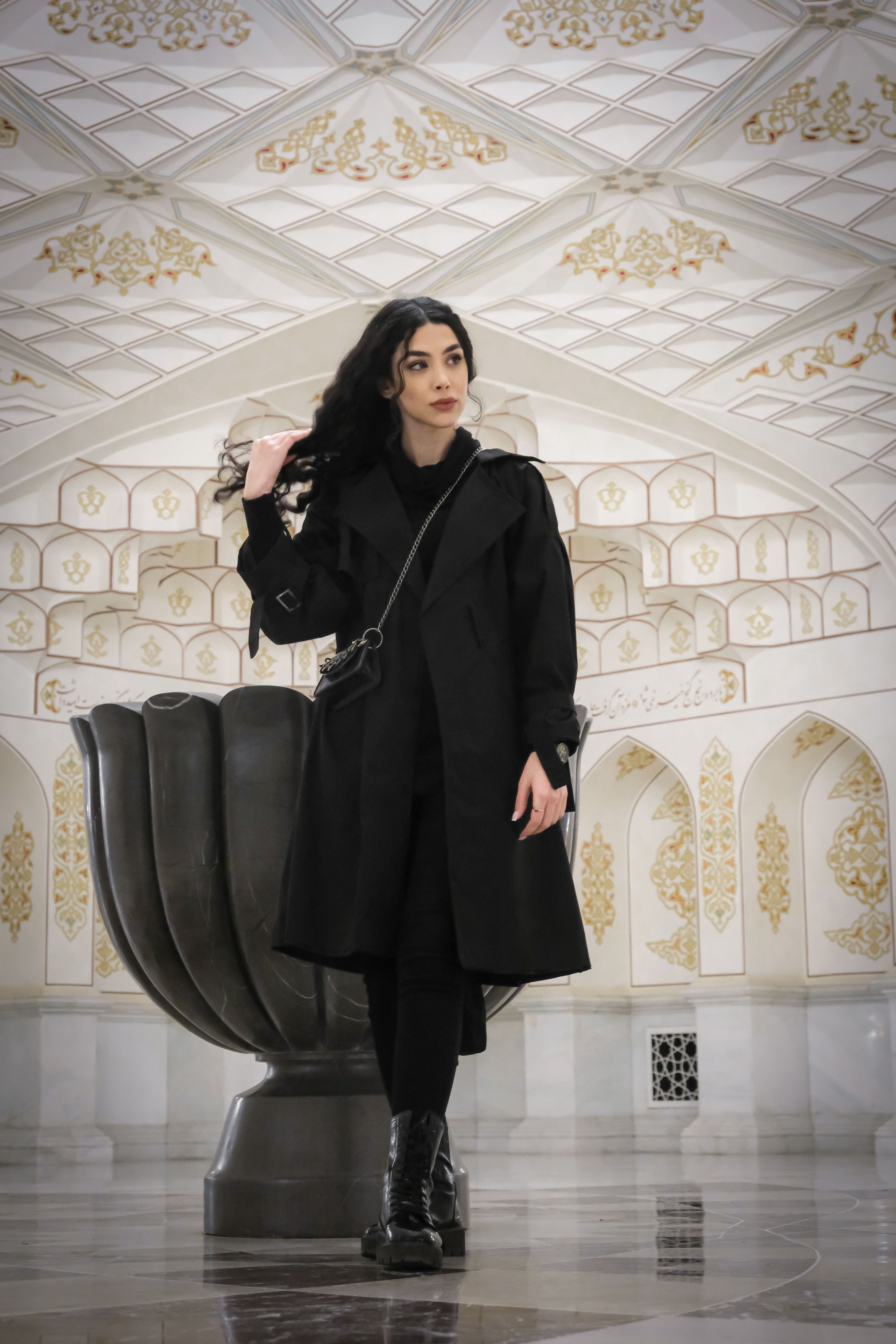 A fashionable woman poses in an ornate indoor setting with detailed architecture.