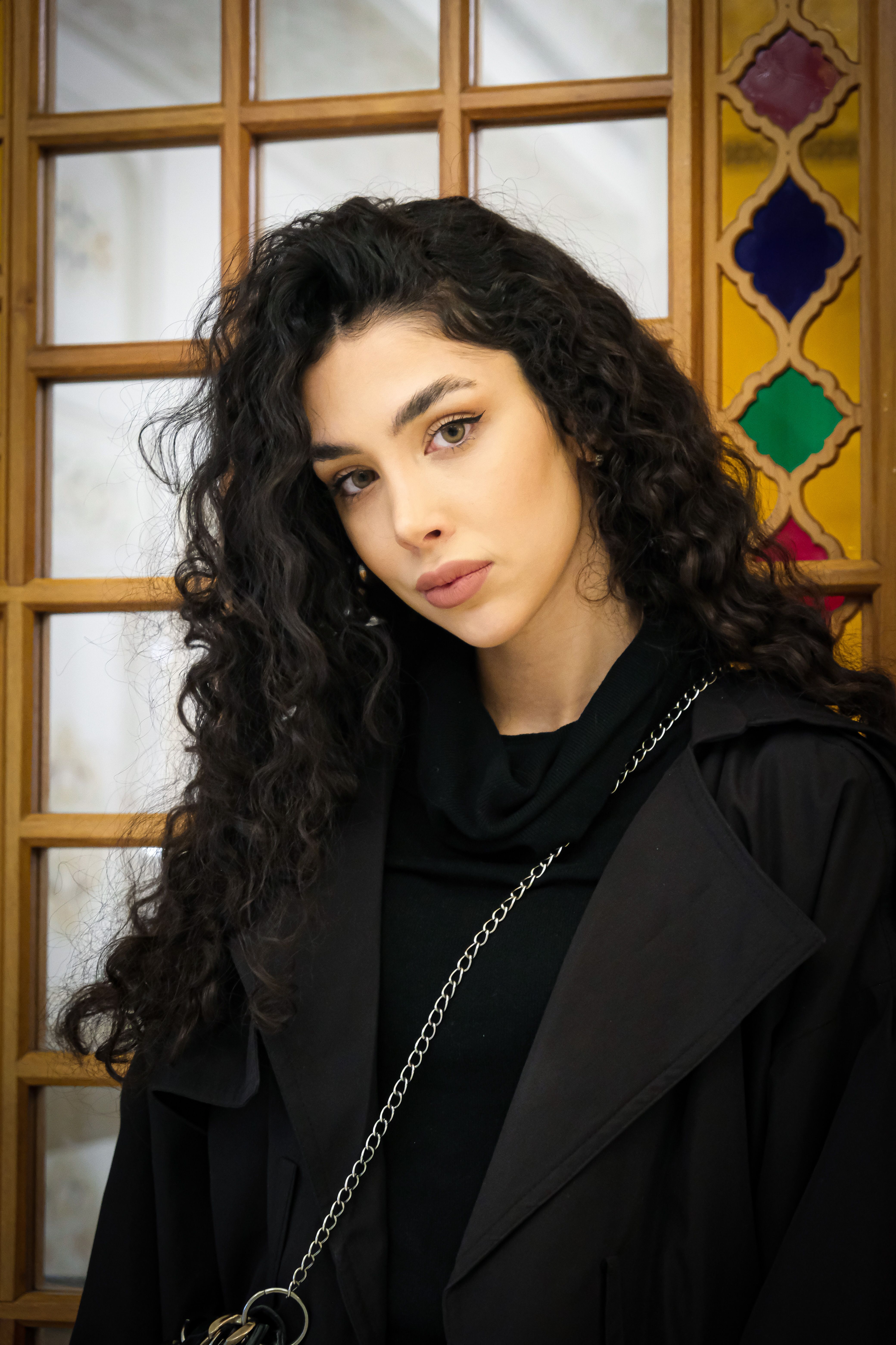 Young woman with curly hair posing by a colorful window indoors.