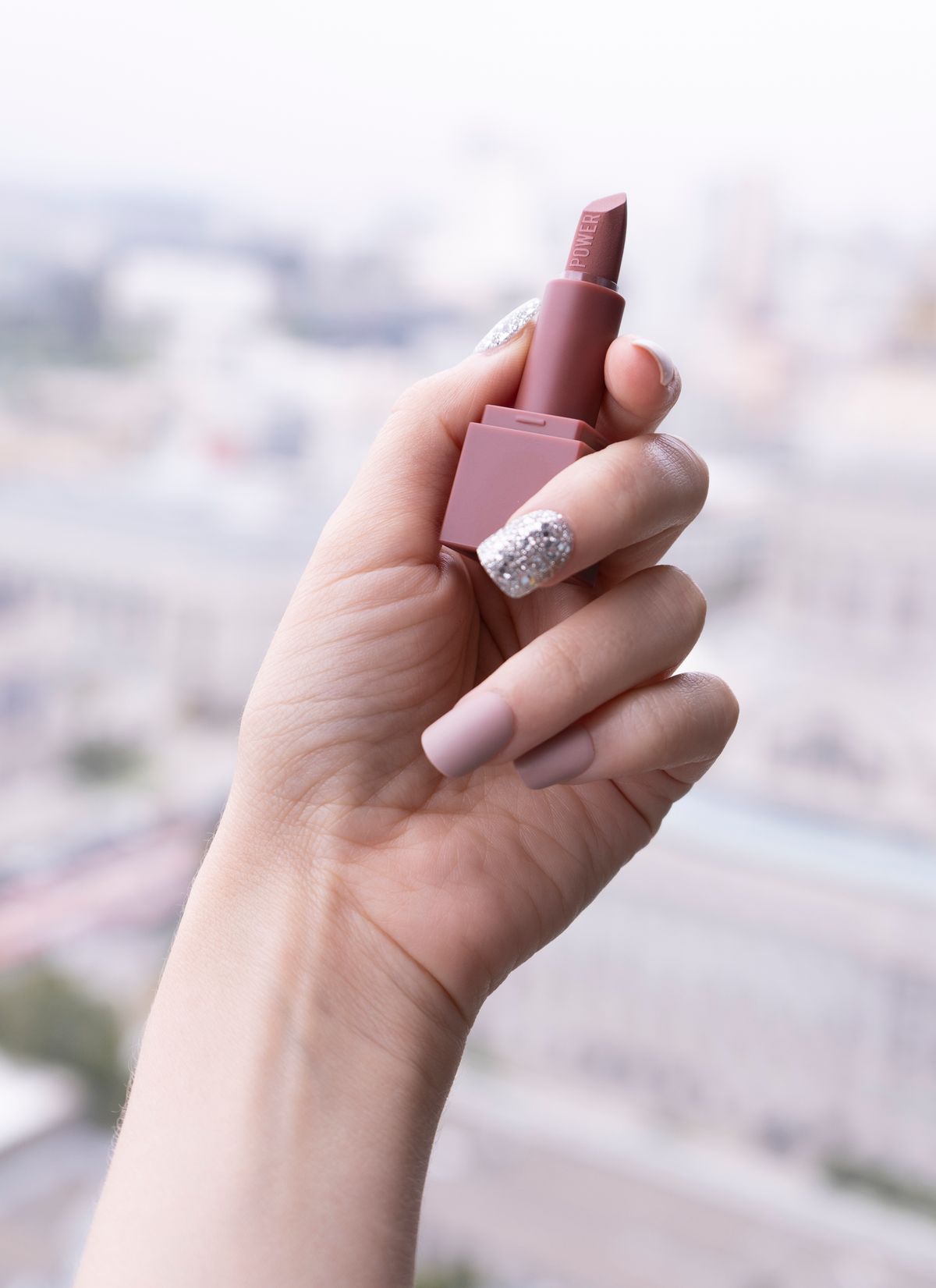Hand with manicured nails holding a nude lipstick with a blurry cityscape in the background.
