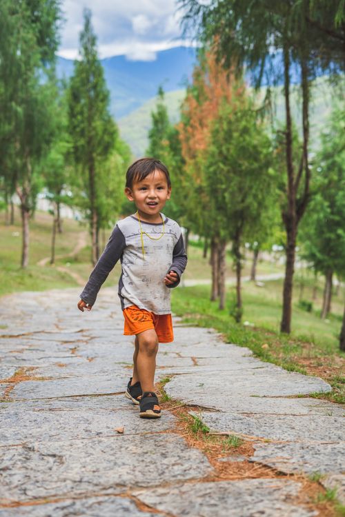 Free A joyful child in orange shorts walks down a tree-lined stone path in a park. Stock Photo