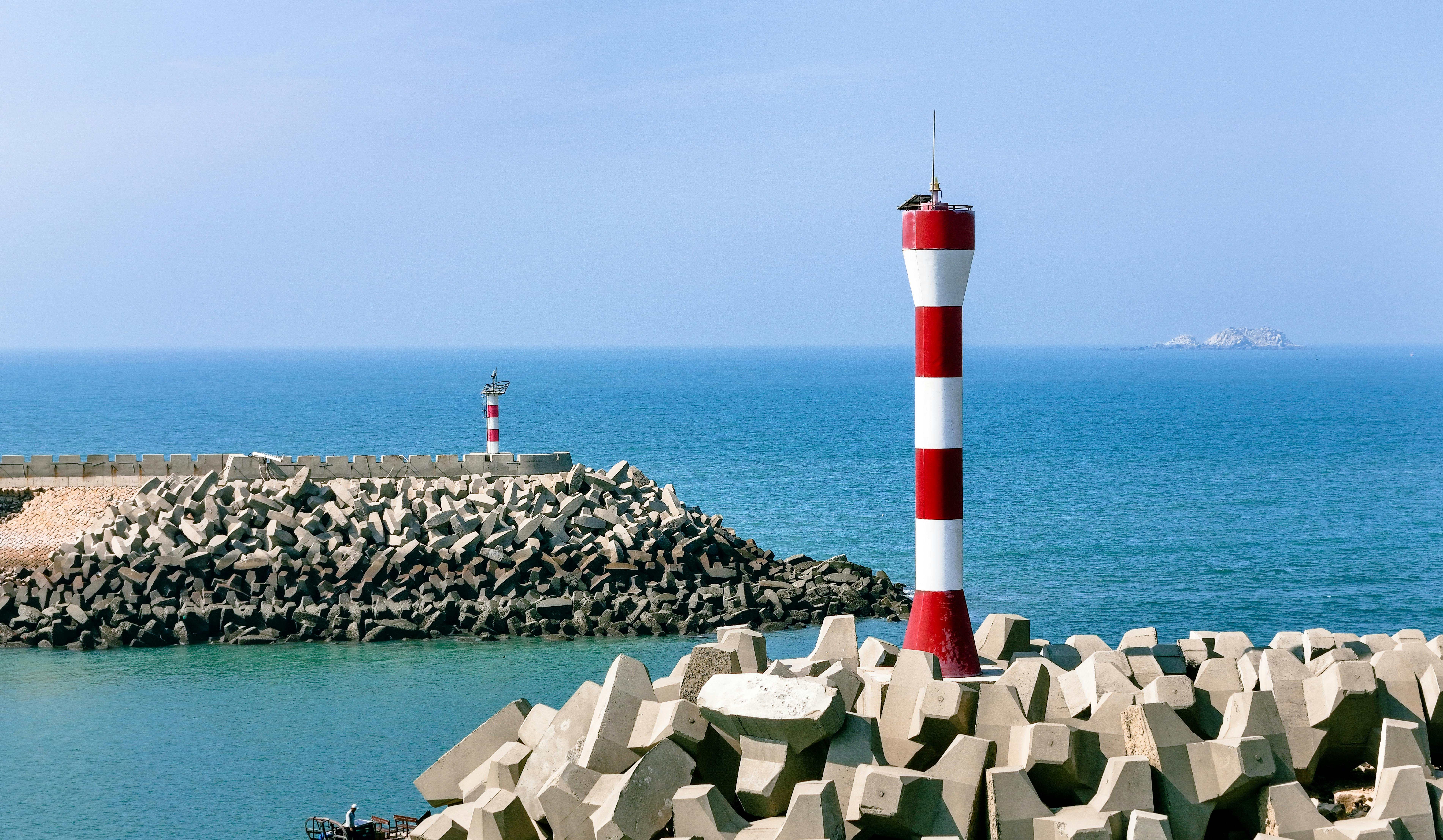 Red and white lighthouse by a rocky seawall against a blue ocean backdrop.