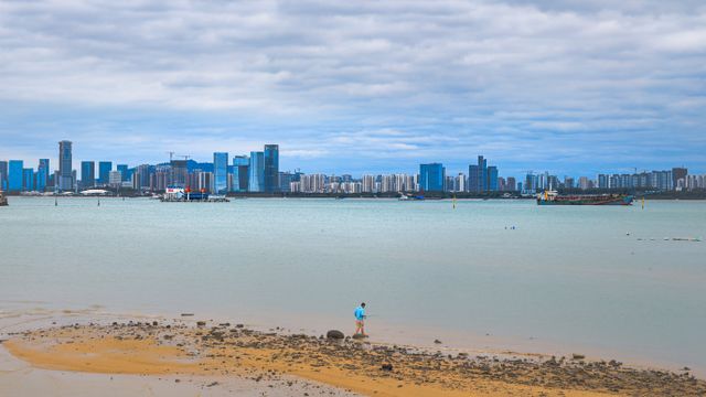 A distant skyline of Haikou, China viewed from a beach, with one person walking on the sand. — lounges at Haikou Airport