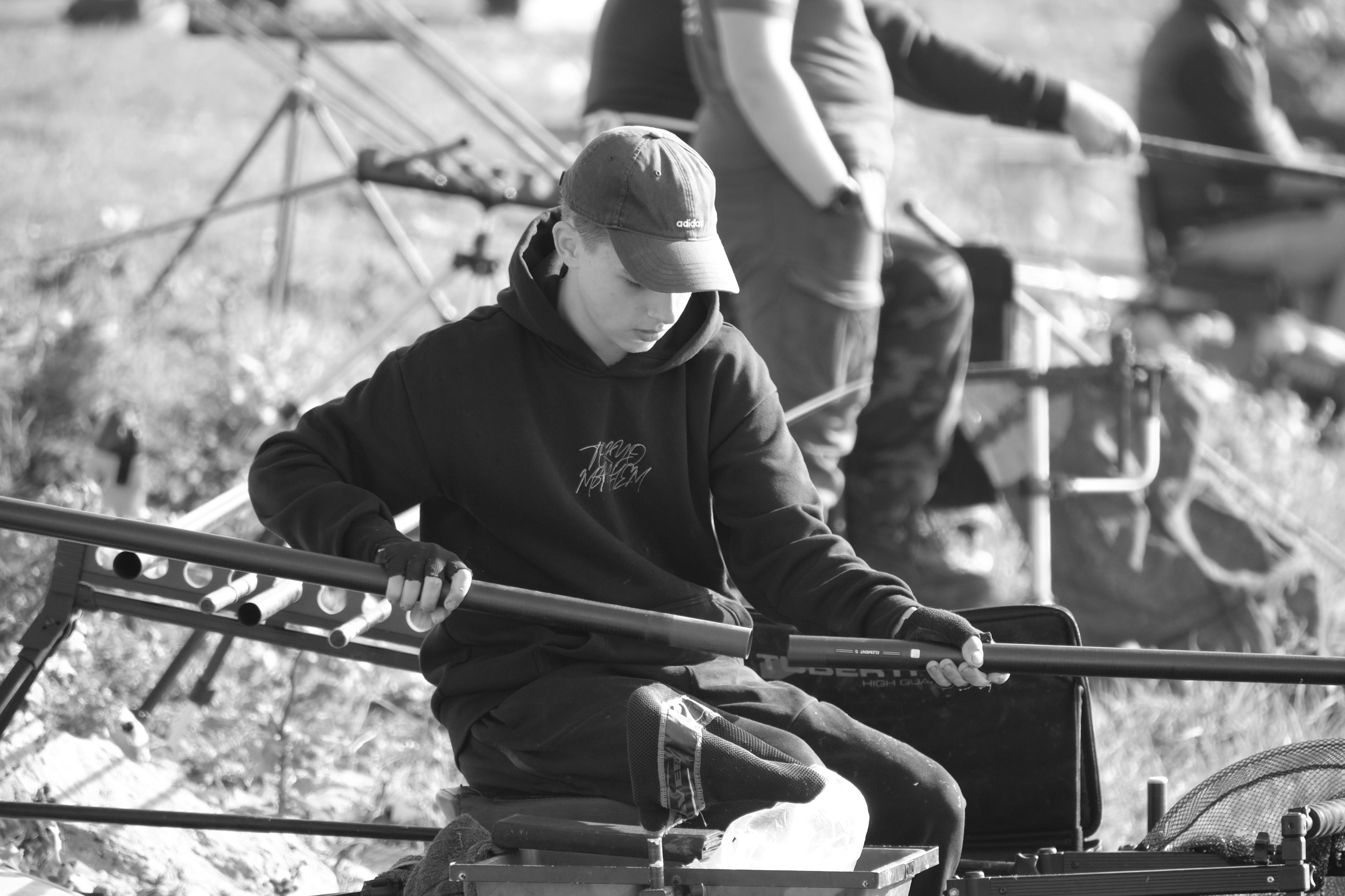 A young man prepares fishing gear by a lakeside in monochrome, capturing the tranquil sport.