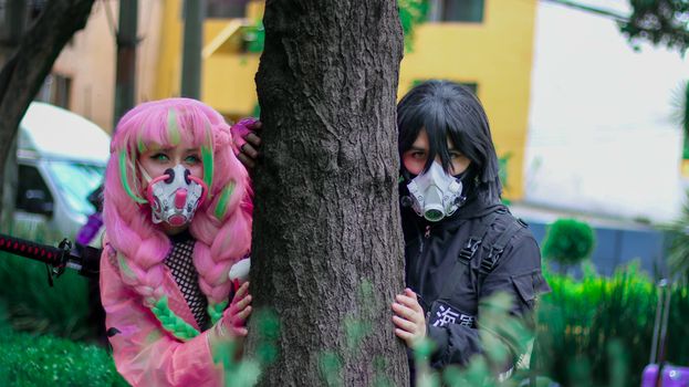 Two people wearing cosplay masks pose by a tree in a city setting.