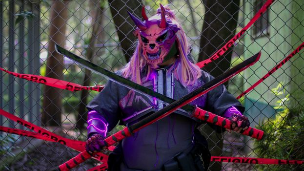 A person in a demon mask holds swords in front of a fence marked with warning tape.