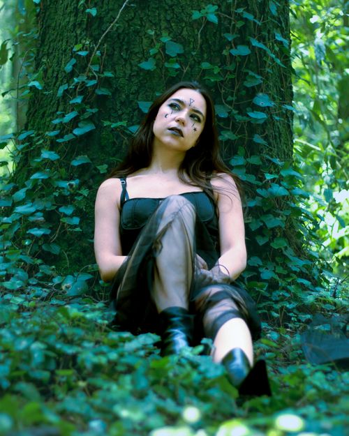 Free A woman in a black dress sits against a tree, surrounded by lush greenery. Stock Photo