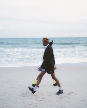 A person in a suit and sneakers walks on a serene beach holding a skateboard.