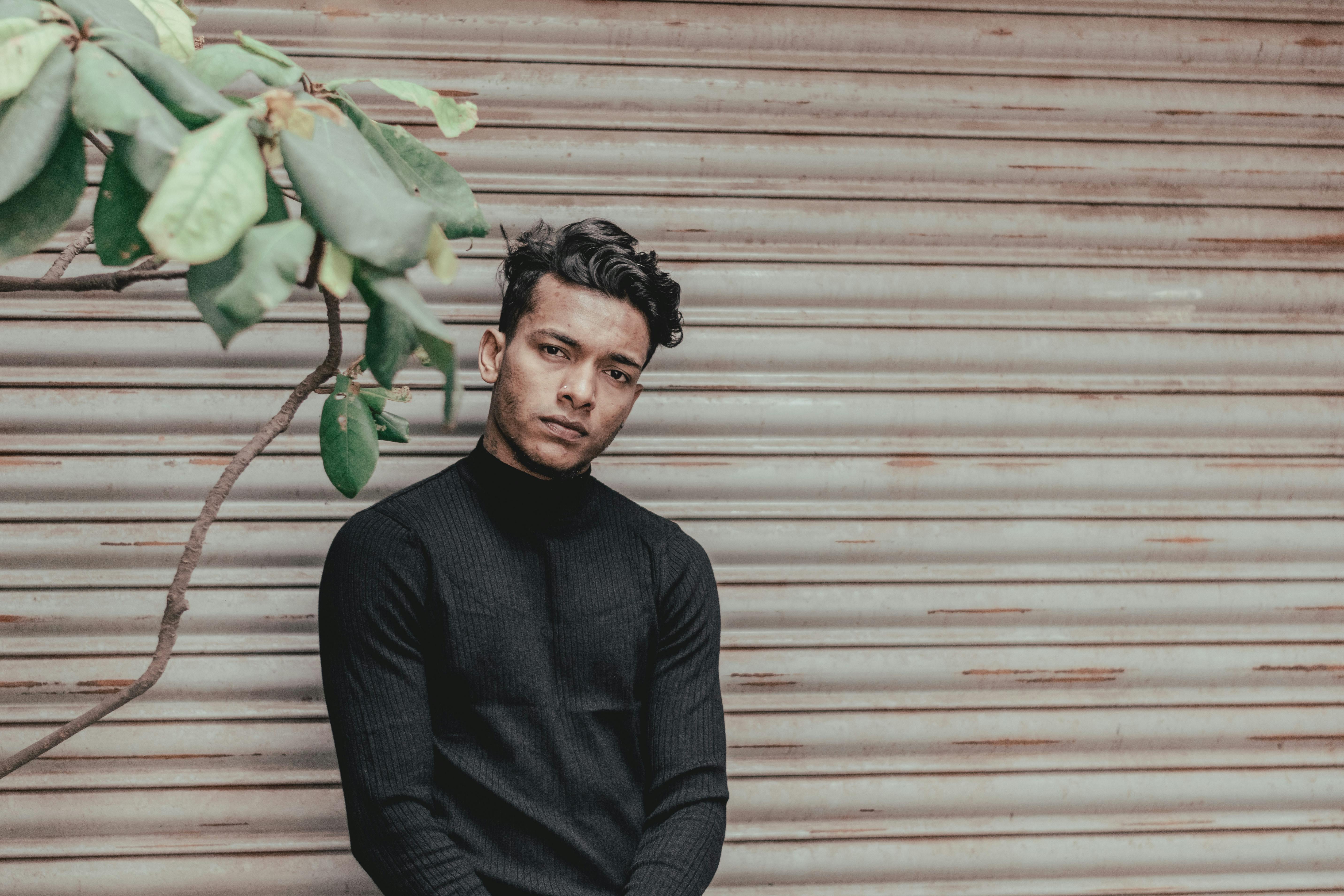 一位时尚男子穿着黑色毛衣，靠在百叶窗旁，旁边有一株植物