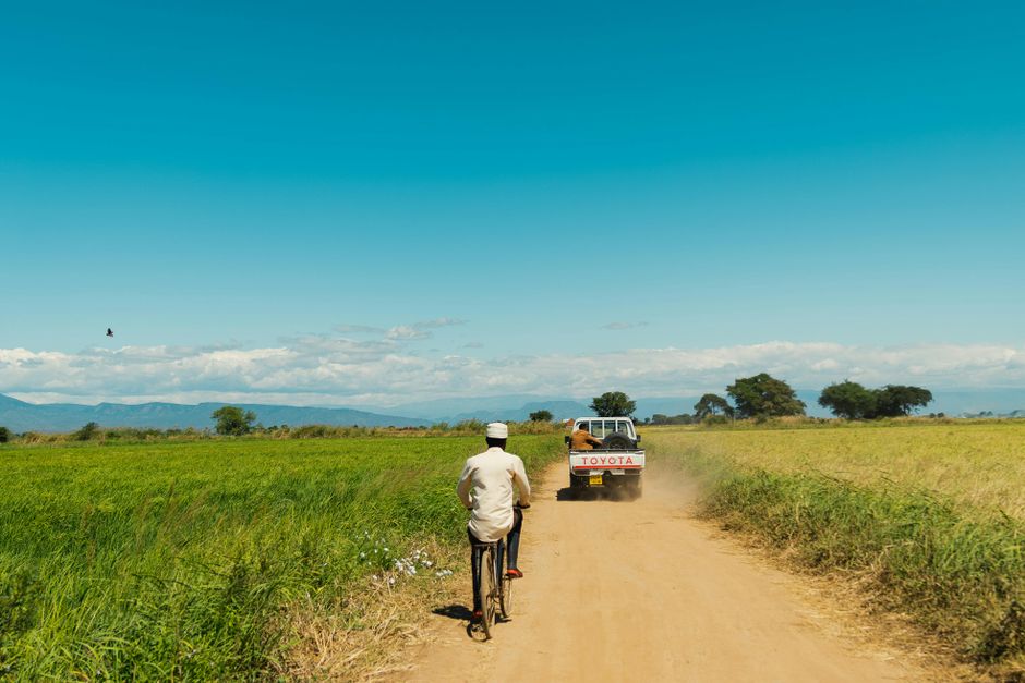 A dirt road in rural Tanzania with a cyclist and a vehicle under a clear blue sky.