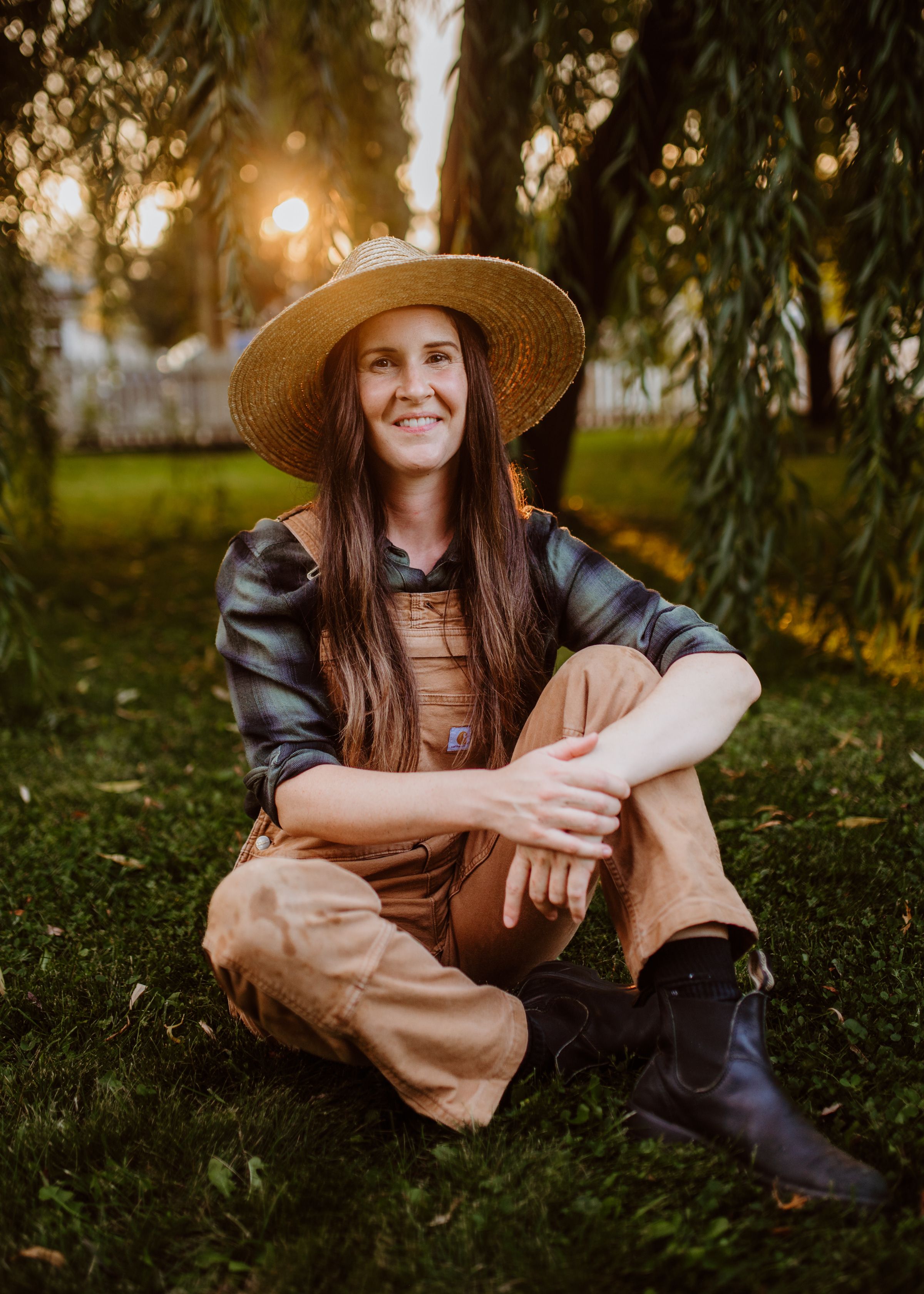 Happy woman gardener sitting on grass wearing overalls and straw hat at sunset.