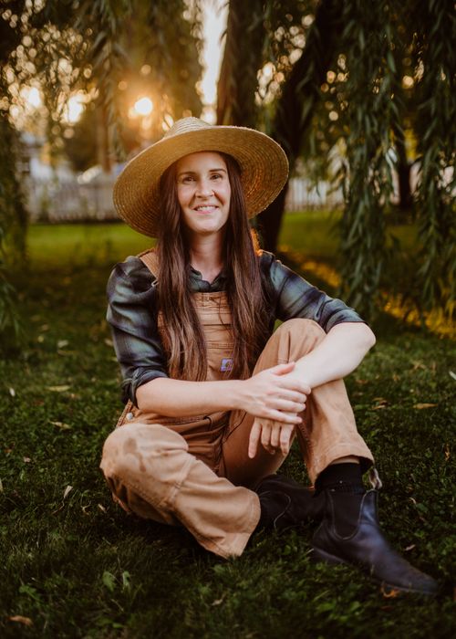 Free Happy woman gardener sitting on grass wearing overalls and straw hat at sunset. Stock Photo