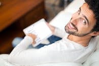 一个快乐的男人坐在家里的沙发上，用数字平板电脑看着相机