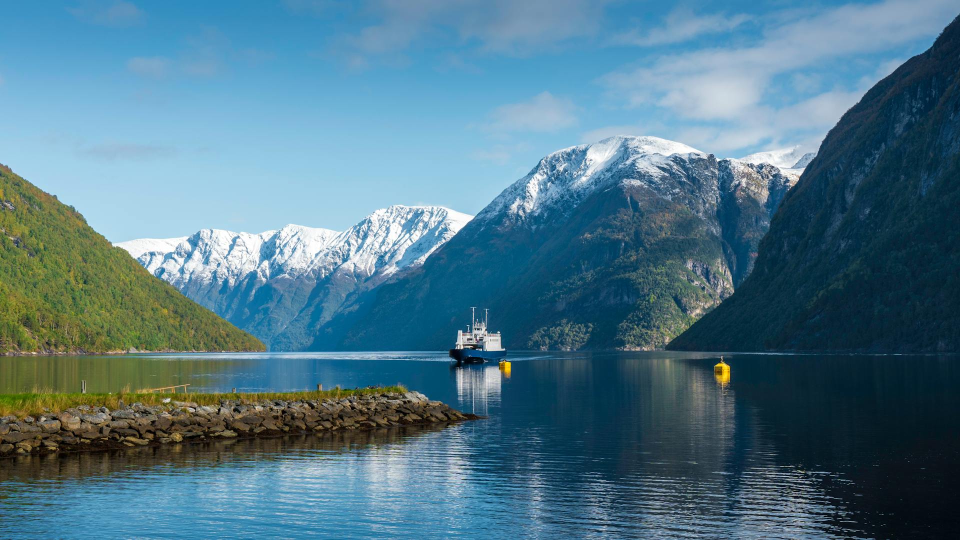 Reflective ferry traversing a stunning Norwegian fjord surrounded by snow-capped mountains.