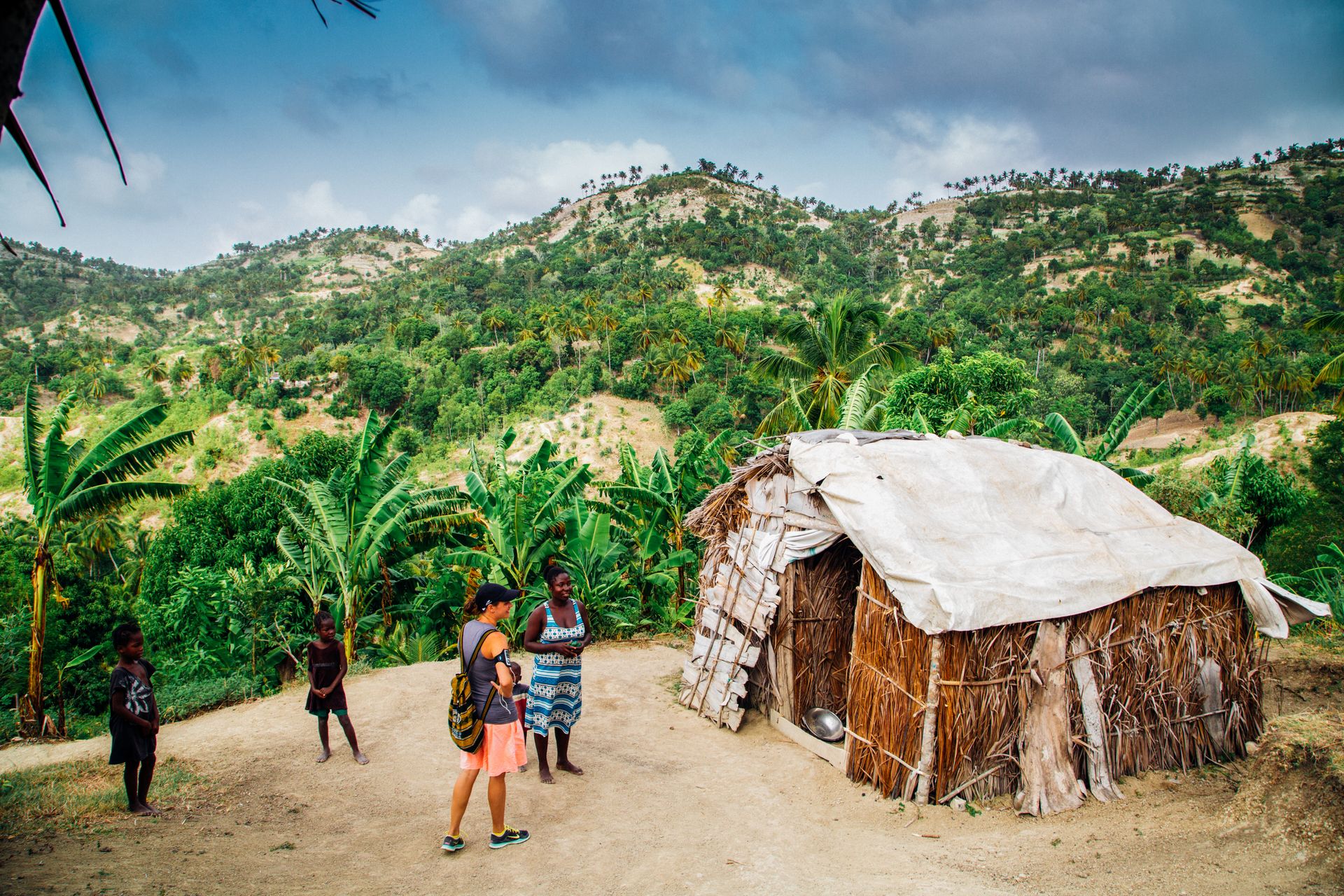 A rural hillside village with people standing near a traditional hut and lush greenery.