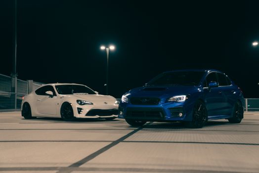 Two sports cars, white and blue, parked on a rooftop at night under streetlights.