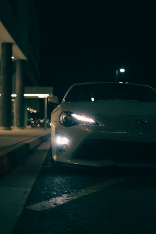 Free White sports car with headlights on, parked along a city street at night, showcasing urban life. Stock Photo