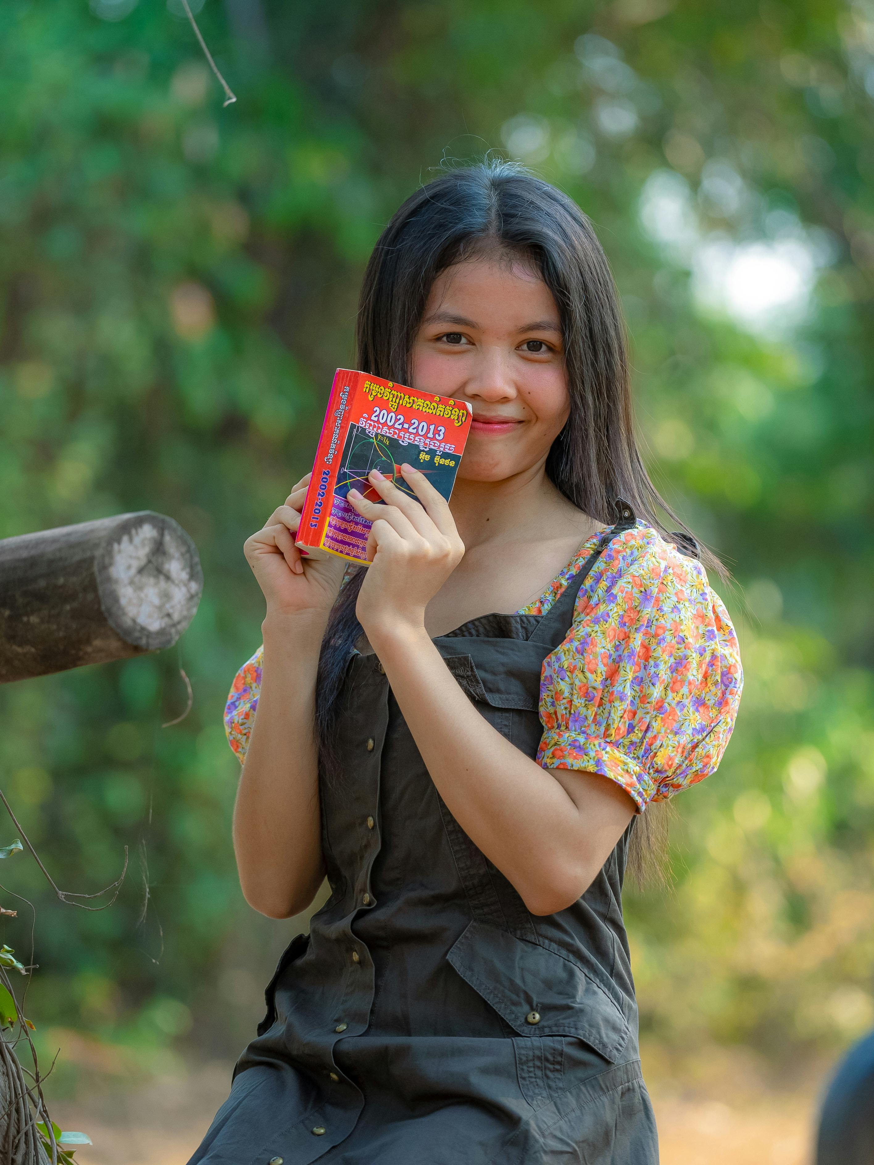 Miễn phí Một cô gái trẻ trong trang phục sặc sỡ đang cầm một cuốn sách trước khung cảnh ngoài trời xanh tươi. Ảnh lưu trữ