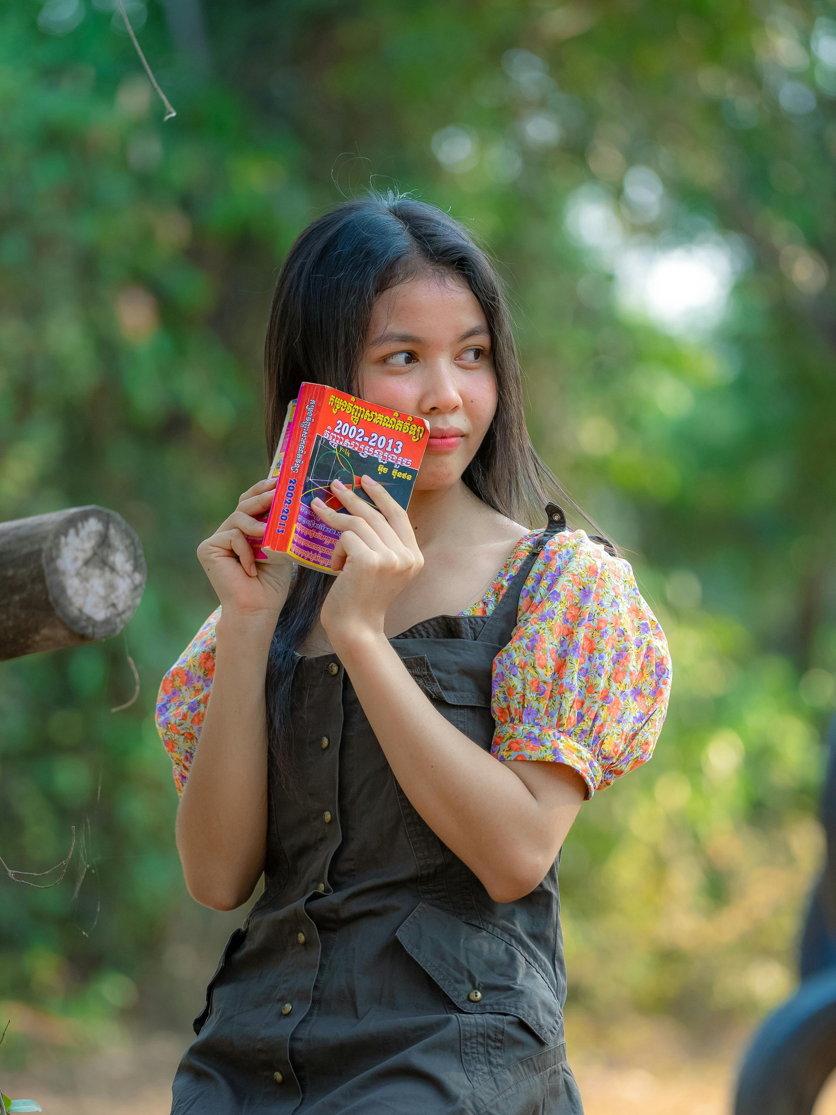 ฟรี หญิงสาวคนหนึ่งโพสท่าถ่ายรูปพร้อมกับอ่านหนังสือ ขณะเพลิดเพลินกับธรรมชาติในบรรยากาศกลางแจ้งที่เงียบสงบ คลังภาพถ่าย