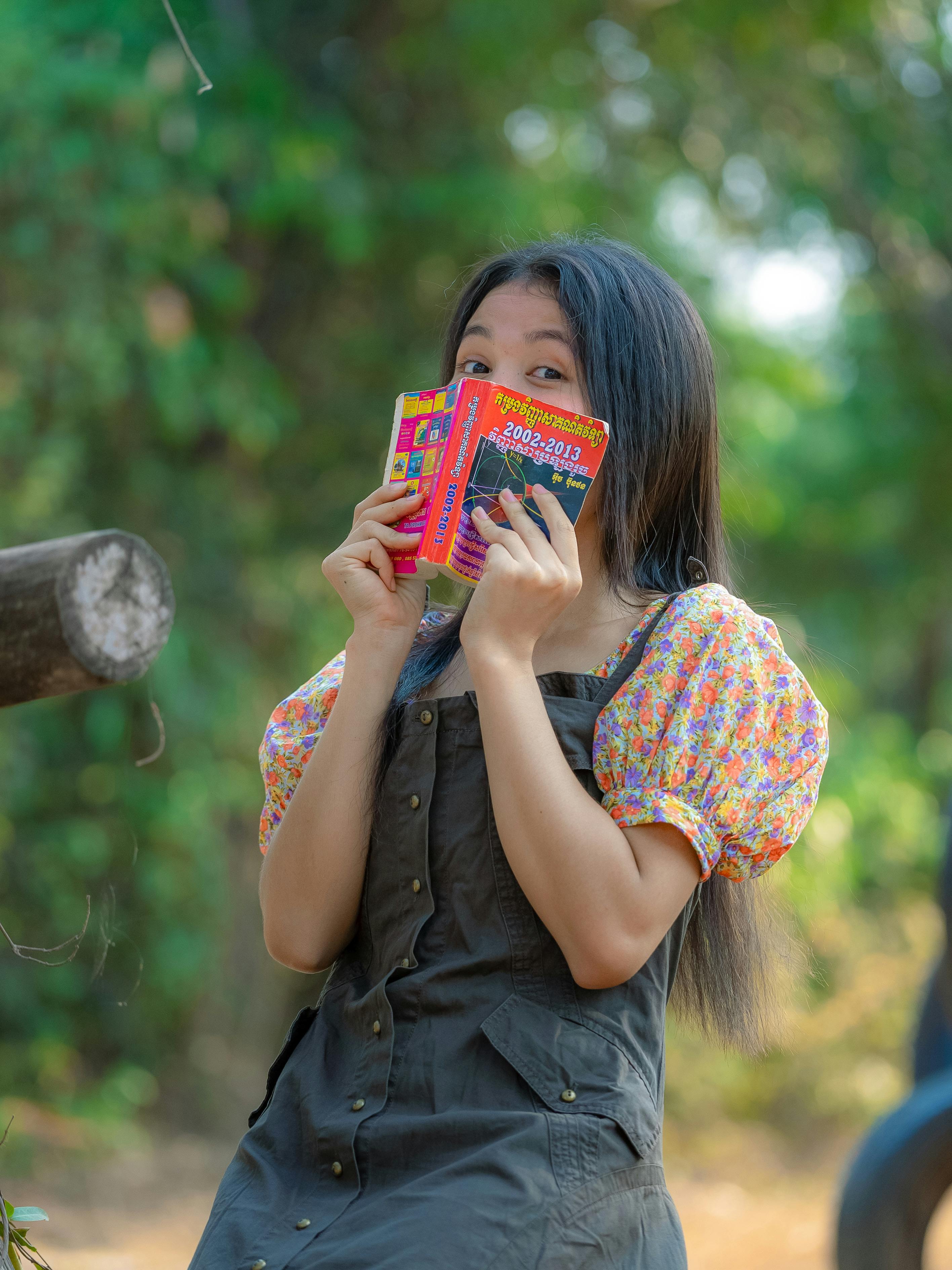 免費 一位年輕女子在花園裡享受陽光明媚的一天時，頑皮地躲在書後面。 圖庫相片